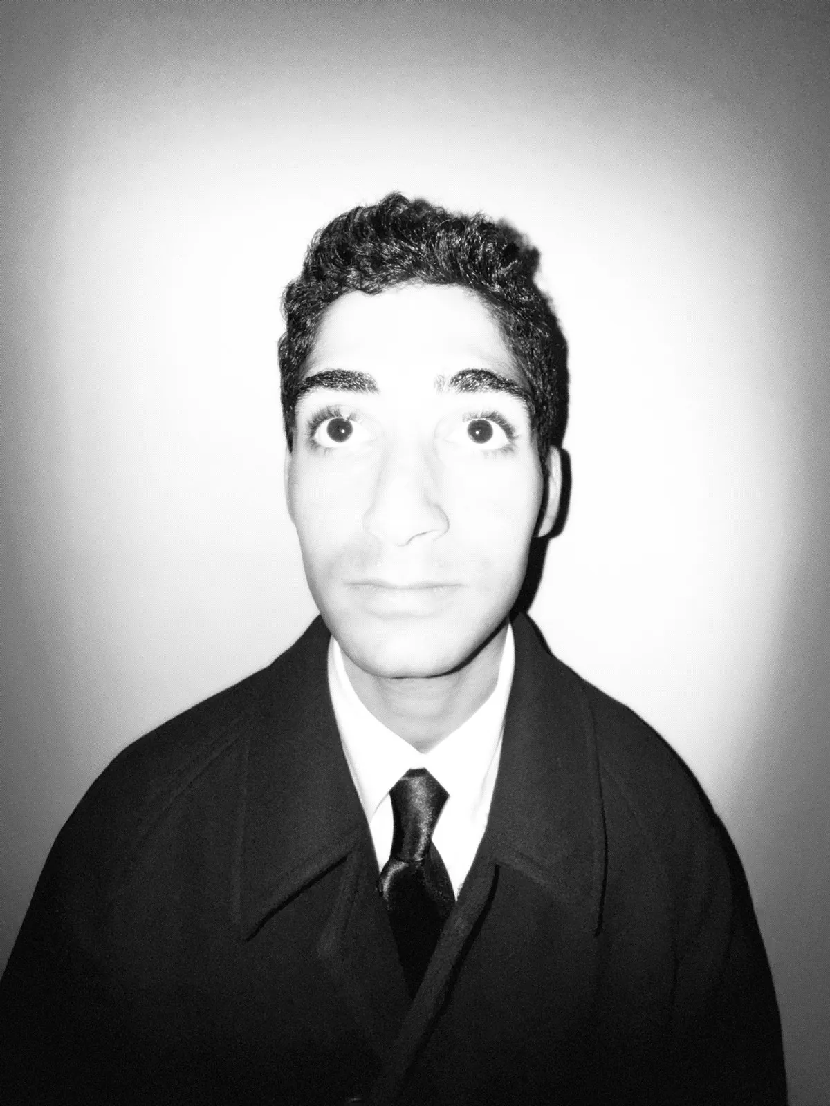 A black-and-white portrait of Mo Badawi in a dark coat and tie, lit softly against a pale gray backdrop.